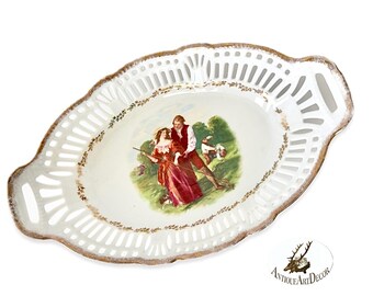 May include: White oval serving dish with a cut-out design and gold trim. The center features a painted scene of a couple in a field with a dog.