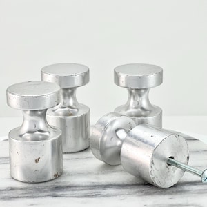 May include: Four silver-colored cabinet knobs with a cylindrical base and a flat, round top. One knob is lying on its side, revealing a screw. The knobs are made of metal and have a slightly worn appearance. The knobs are designed for furniture.