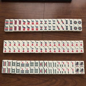 May include: A set of ivory-colored Mahjong tiles arranged on a wooden surface. The tiles feature various symbols, numbers, and characters in red, green, and blue. The game pieces are organized in rows, showcasing the different tile designs.