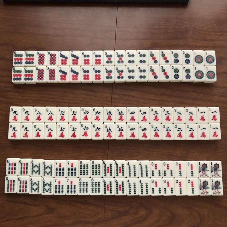 May include: A set of ivory-colored Mahjong tiles arranged on a wooden surface. The tiles feature various symbols, numbers, and characters in red, green, and blue. The game pieces are organized in rows, showcasing the different tile designs.