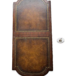 May include: A two-part, brown leather desk blotter with gold trim. The blotter has a rectangular shape with a curved top and bottom. The leather has a rich, aged appearance, and the gold trim adds a touch of elegance.