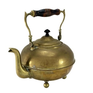 May include: Antique brass teapot with a wooden handle and three feet. The teapot has a rounded body and a lid with a knob.