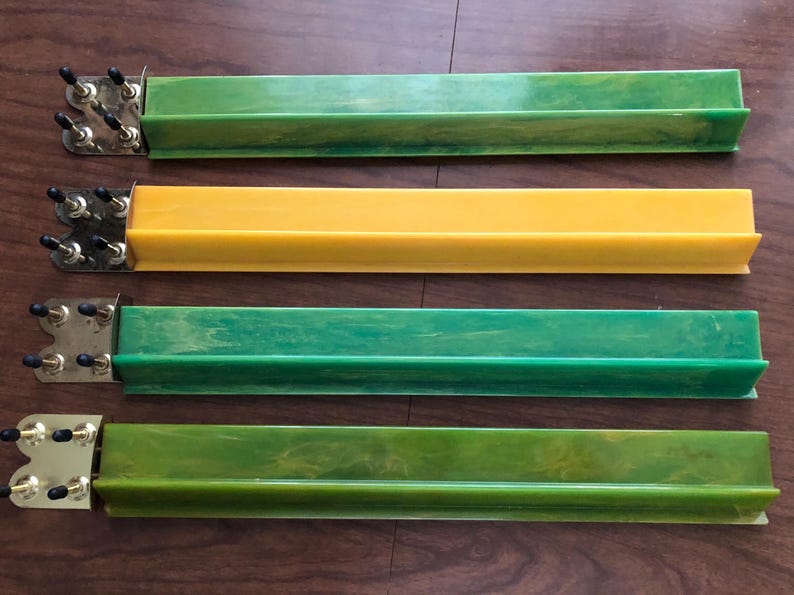 May include: Four vintage Mahjong tile racks in shades of green and yellow. Each rack has a rectangular trough design with a metal end piece featuring black knobs. The racks are arranged horizontally on a wooden surface.