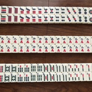 May include: Three rows of ivory-colored Mahjong tiles are arranged on a wooden surface. The tiles display various symbols and numbers in red, green, and black. The game pieces are rectangular and feature intricate designs.
