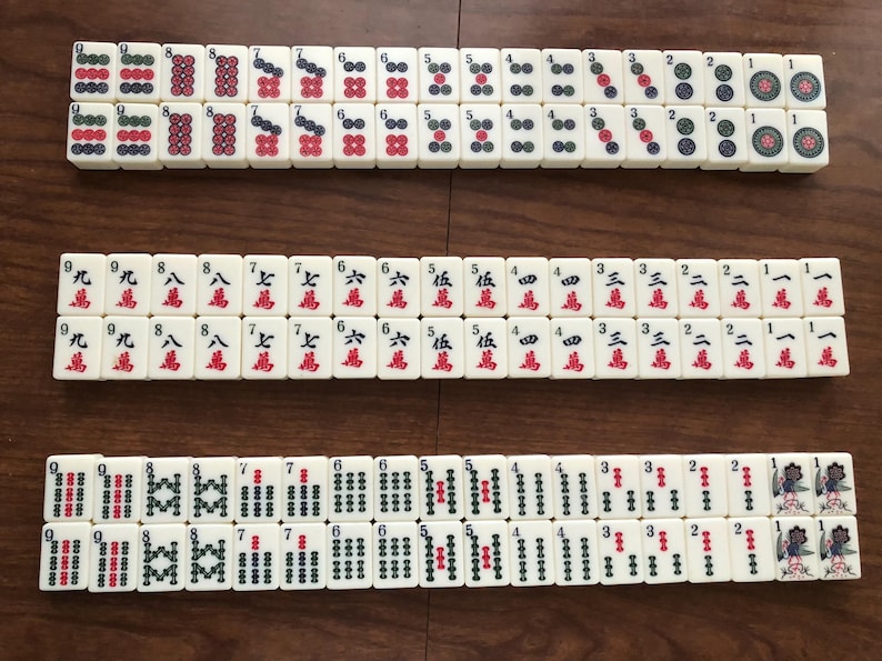 May include: Three rows of ivory-colored Mahjong tiles are arranged on a wooden surface. The tiles display various symbols and numbers in red, green, and black. The game pieces are rectangular and feature intricate designs.