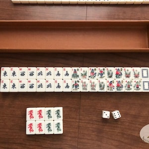 May include: A vintage Mahjong set with ivory-colored tiles, featuring Chinese characters and colorful illustrations. The set includes a brown leatherette case, dice, and a selection of tiles arranged on a wooden surface. The tiles display various symbols and images.
