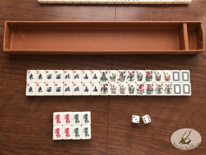 May include: A vintage Mahjong set with ivory-colored tiles, featuring Chinese characters and colorful illustrations. The set includes a brown leatherette case, dice, and a selection of tiles arranged on a wooden surface. The tiles display various symbols and images.