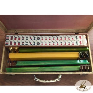 May include: Open wooden mahjong set with tiles, racks, and accessories. The tiles are white with red and green markings. The set includes green and yellow racks, and is stored in a wooden case with a handle.