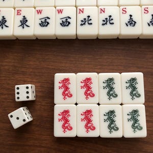 May include: A collection of ivory-colored Mahjong tiles with black, red, and green symbols and characters. Two white dice with black dots are also present. The tiles are arranged on a brown wooden surface.