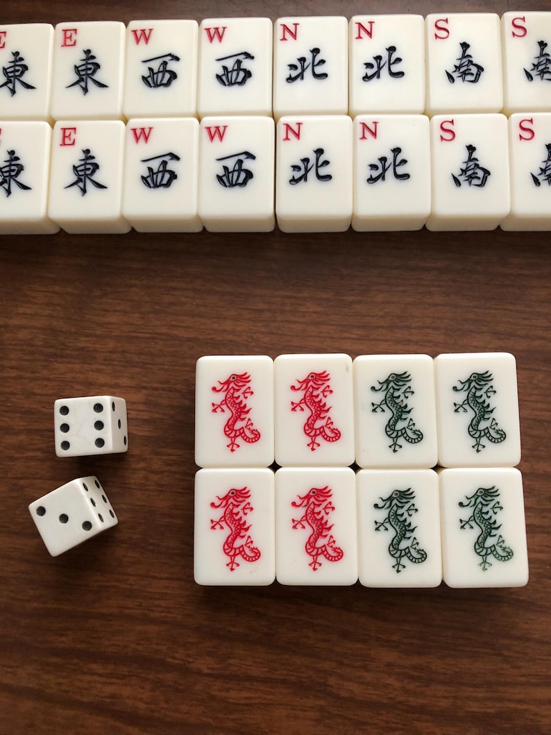 May include: A collection of ivory-colored Mahjong tiles with black, red, and green symbols and characters. Two white dice with black dots are also present. The tiles are arranged on a brown wooden surface.