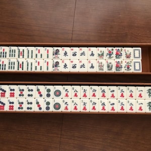 May include: A complete Mahjong set with ivory-colored tiles arranged in two wooden trays. The tiles feature Chinese characters and symbols in red, green, and black. Two dice are visible in one tray. The game is set on a brown surface.