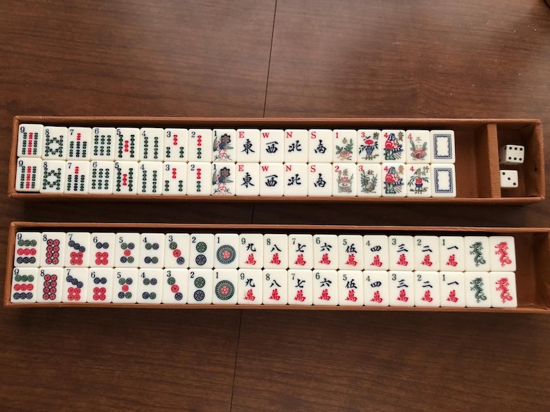 May include: A complete Mahjong set with ivory-colored tiles arranged in two wooden trays. The tiles feature Chinese characters and symbols in red, green, and black. Two dice are visible in one tray. The game is set on a brown surface.