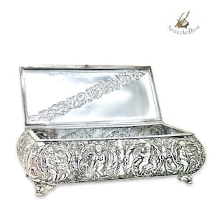 May include: A silver-toned antique box with an open lid. The box features detailed relief carvings of figures and foliage around the sides. The lid's interior has an ornate floral design. The box rests on four small feet.