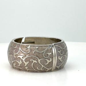 May include: A silver-tone hinged bracelet with a light pink enamel finish. The bracelet features a scrolling, floral design in a lighter silver color. The bracelet is a wide cuff style.