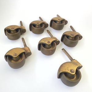 May include: A collection of seven brass furniture casters, each featuring a rounded wheel housing and a threaded metal stem for attachment. The brass components exhibit a muted, metallic finish with visible signs of age and patina throughout.