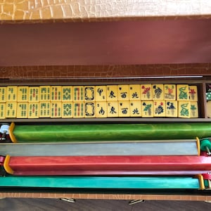 May include: Open brown leatherette case containing a complete Mahjong set. The set includes yellow tiles with black symbols, dice, and colorful racks. The case is lined with a lighter brown material.