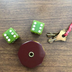 Puede incluir: Un surtido de piezas de juego vintage que incluye tres dados verdes con puntos blancos, una pieza octogonal granate y amarilla, y una peque&ntilde;a llave de lat&oacute;n con una cuerda roja. Los art&iacute;culos est&aacute;n dispuestos sobre una superficie de madera.