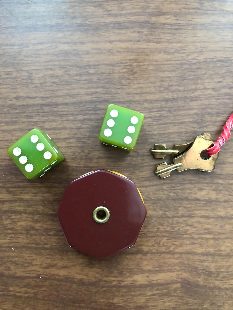 Puede incluir: Un surtido de piezas de juego vintage que incluye tres dados verdes con puntos blancos, una pieza octogonal granate y amarilla, y una peque&ntilde;a llave de lat&oacute;n con una cuerda roja. Los art&iacute;culos est&aacute;n dispuestos sobre una superficie de madera.