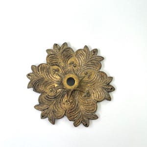 May include: A circular, antique brass-colored metal decorative plate featuring a detailed, embossed floral leaf pattern radiating from a central circular opening. The piece has a weathered, textured finish with small holes along the outer edges.