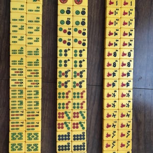 Puede incluir: Tres columnas de fichas de Mahjong amarillas est&aacute;n dispuestas sobre una superficie marr&oacute;n. Las fichas muestran varios s&iacute;mbolos, n&uacute;meros y caracteres en rojo, verde y negro. Las fichas son rectangulares y tienen esquinas redondeadas, con un acabado liso y brillante.