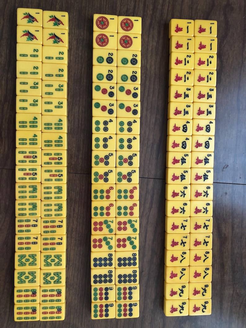 Puede incluir: Tres columnas de fichas de Mahjong amarillas est&aacute;n dispuestas sobre una superficie marr&oacute;n. Las fichas muestran varios s&iacute;mbolos, n&uacute;meros y caracteres en rojo, verde y negro. Las fichas son rectangulares y tienen esquinas redondeadas, con un acabado liso y brillante.