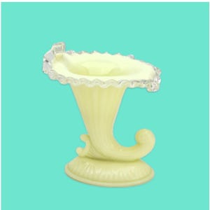 May include: A pale yellow, vintage-style glass vase with a ruffled rim and a decorative, curved base. The vase has a flared, trumpet-like shape with a clear, scalloped edge. The base features a swirl design, adding to its ornate appearance.