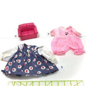 May include: A collection of doll accessories, including a pink miniature sofa, a blue and white strawberry print dress, and a pink romper with a floral applique. The items are arranged on a white surface.