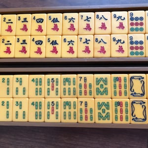 Puede incluir: Un juego de Mahjong vintage con fichas de color marfil, cada una con caracteres y s&iacute;mbolos chinos en rojo, verde y azul. Las fichas est&aacute;n dispuestas en bandejas de madera, mostrando los componentes del juego.