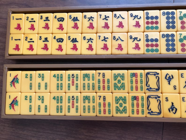 Puede incluir: Un juego de Mahjong vintage con fichas de color marfil, cada una con caracteres y s&iacute;mbolos chinos en rojo, verde y azul. Las fichas est&aacute;n dispuestas en bandejas de madera, mostrando los componentes del juego.