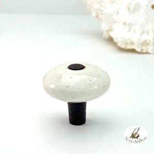 May include: A bottle stopper featuring a smooth, off-white, rounded ceramic top with a dark brown center accent. The base is a dark, cylindrical material designed for insertion into a bottle neck, providing a functional seal for various glass containers.