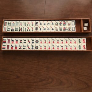 May include: A vintage Mahjong set with ivory-colored tiles featuring Chinese characters and symbols in red, green, and blue. The set includes dice and is stored in two wooden trays.