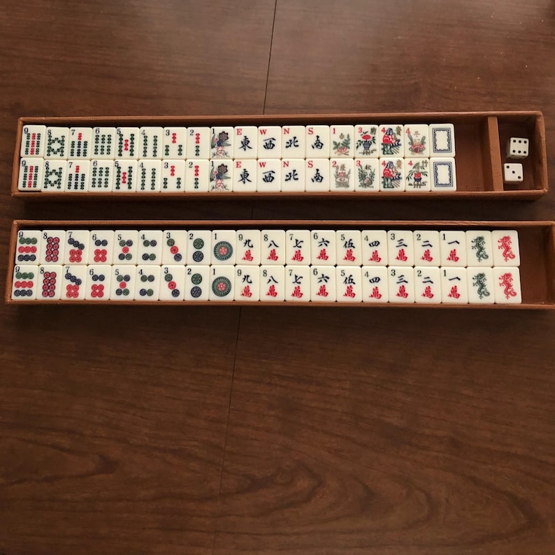 May include: A vintage Mahjong set with ivory-colored tiles featuring Chinese characters and symbols in red, green, and blue. The set includes dice and is stored in two wooden trays.