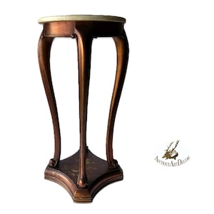 May include: A bronze-colored plant stand with a round, cream-colored top. The stand has three curved legs and a decorative base. The overall design is ornate and antique-inspired, suitable for displaying plants or decorative items.