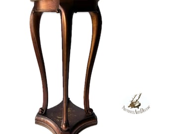 May include: A bronze-colored plant stand with a round, cream-colored top. The stand has three curved legs and a decorative base. The overall design is ornate and antique-inspired, suitable for displaying plants or decorative items.