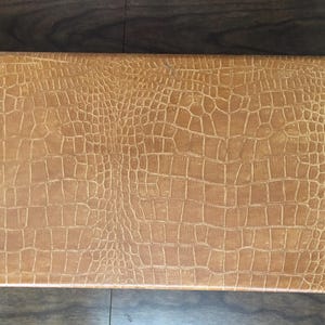 Puede incluir: Una caja rectangular de color marr&oacute;n claro con un patr&oacute;n texturizado de piel de cocodrilo. La caja est&aacute; cerrada y parece estar hecha de cuero o un material similar. La caja est&aacute; sobre una superficie de madera oscura.