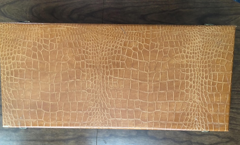Puede incluir: Una caja rectangular de color marr&oacute;n claro con un patr&oacute;n texturizado de piel de cocodrilo. La caja est&aacute; cerrada y parece estar hecha de cuero o un material similar. La caja est&aacute; sobre una superficie de madera oscura.
