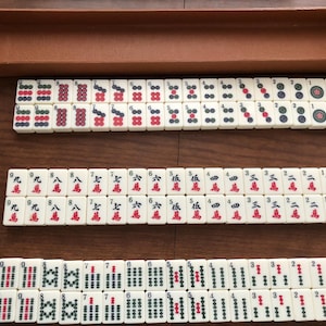May include: A complete Mahjong set with ivory-colored tiles, featuring red, green, and black symbols and characters. The set includes a brown wooden box for storage, and the tiles are arranged in rows on a wooden surface.