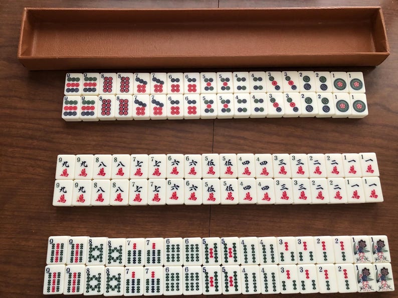 May include: A complete Mahjong set with ivory-colored tiles, featuring red, green, and black symbols and characters. The set includes a brown wooden box for storage, and the tiles are arranged in rows on a wooden surface.