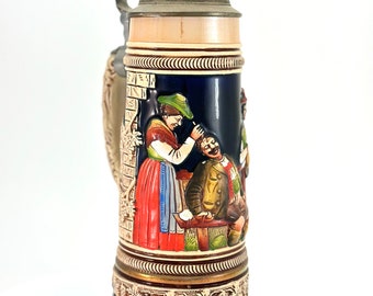 May include: A ceramic beer stein with a blue and gold design. The stein features a scene of three people in traditional German clothing. The lid is made of metal and has a decorative design.
