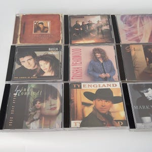 May include: A collection of compact discs featuring country music artists. The CDs showcase album covers with various artists, including Vince Gill, Trisha Yearwood, and Linda Ronstadt. The covers display portraits and album titles.