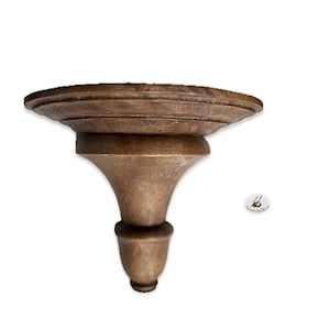 May include: A brown, wooden wall shelf with a decorative, turned design. The shelf has a circular top and a flared, pedestal-style support. The wood has a warm, natural finish, and the shelf is designed for mounting on a wall.