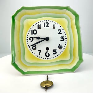 May include: A square, ceramic clock with a white clock face and black numbers. The clock face is surrounded by concentric layers of yellow, green, and white. A gold-colored pendulum hangs below the clock.