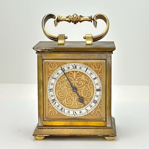 May include: A gold-toned brass mantel clock with a decorative, ornate face and Roman numeral markings. The clock has a handle on the top and sits on four small feet.
