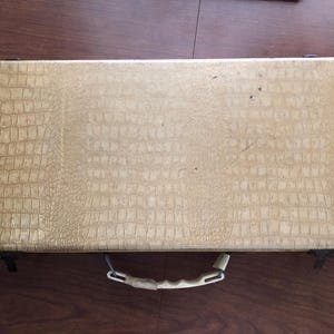 May include: A rectangular, beige, faux-leather case with a crocodile skin pattern. The case has a white handle and black metal clasps. The case is closed and appears to be empty. The case is sitting on a wooden surface.