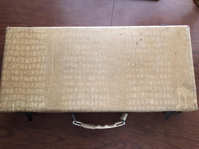 May include: A rectangular, beige, faux-leather case with a crocodile skin pattern. The case has a white handle and black metal clasps. The case is closed and appears to be empty. The case is sitting on a wooden surface.