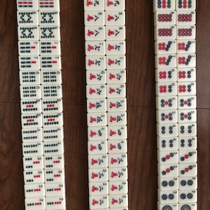 May include: Three columns of ivory-colored Mahjong tiles arranged on a wooden surface. The tiles feature various symbols, numbers, and characters in red, green, and black. The game pieces are neatly stacked, showcasing the intricate designs.