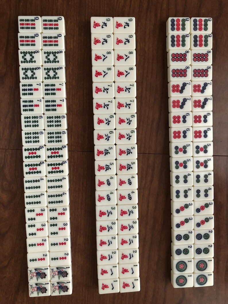 May include: Three columns of ivory-colored Mahjong tiles arranged on a wooden surface. The tiles feature various symbols, numbers, and characters in red, green, and black. The game pieces are neatly stacked, showcasing the intricate designs.
