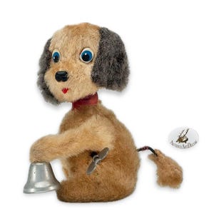 May include: A vintage wind-up dog toy with a bell. The plush dog has tan and gray fur, blue eyes, and a red collar. It is holding a silver bell and has a wind-up key on its side. The tail is brown and black.