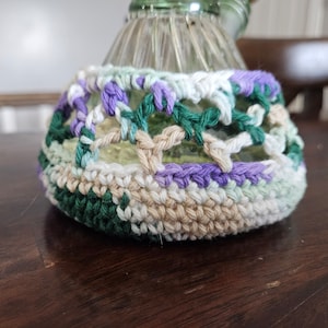 May include: A crocheted bottle cover in shades of green, white, beige, and purple. The cover has a lacy design and is made with a variety of stitches.