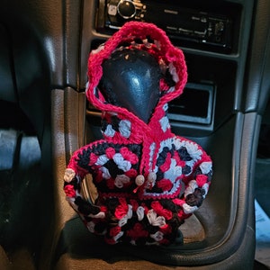 May include: A crocheted car seat cover in black, white and pink. The cover is shaped like a hooded sweater and has a crocheted pattern.